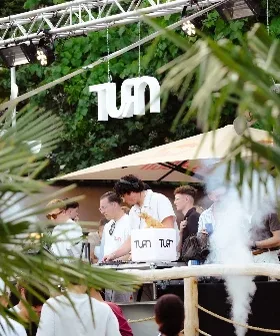 DJ-Pult unter der TURN-Leuchtschrift am Nachmittag, zwischen Palmen und Sonnenschirmen